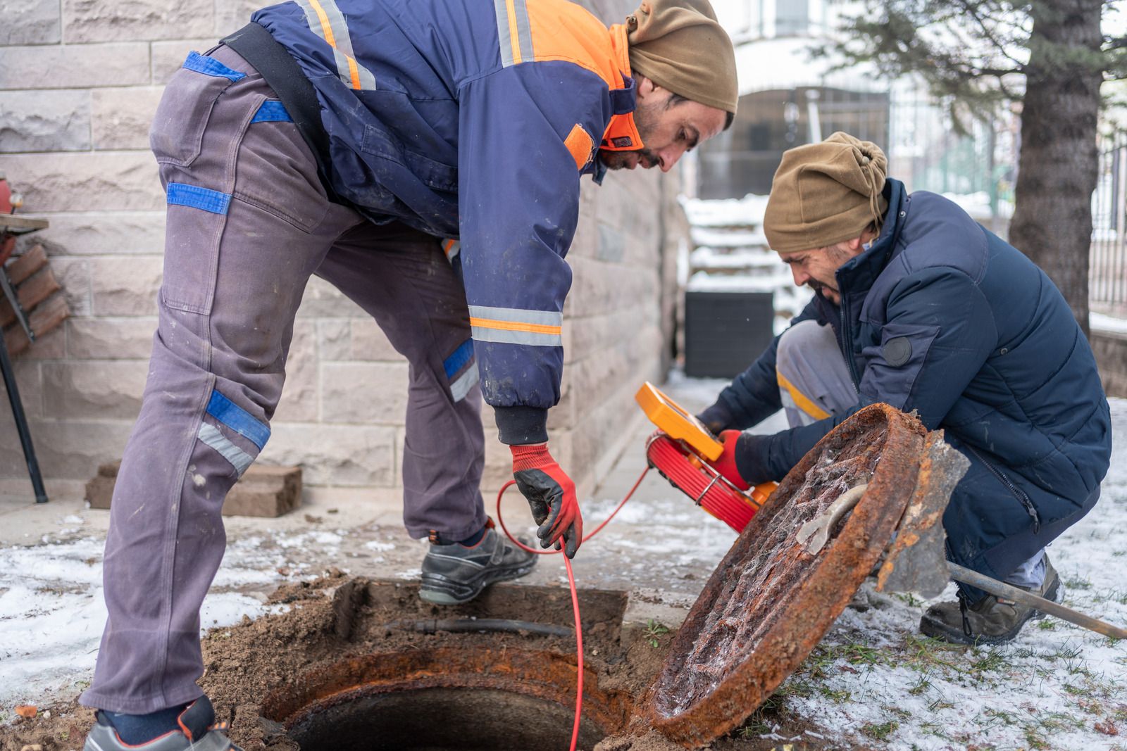 To kloakarbejdere i varmt arbejdstøj undersøger en brønd med inspektionskamera og skærm