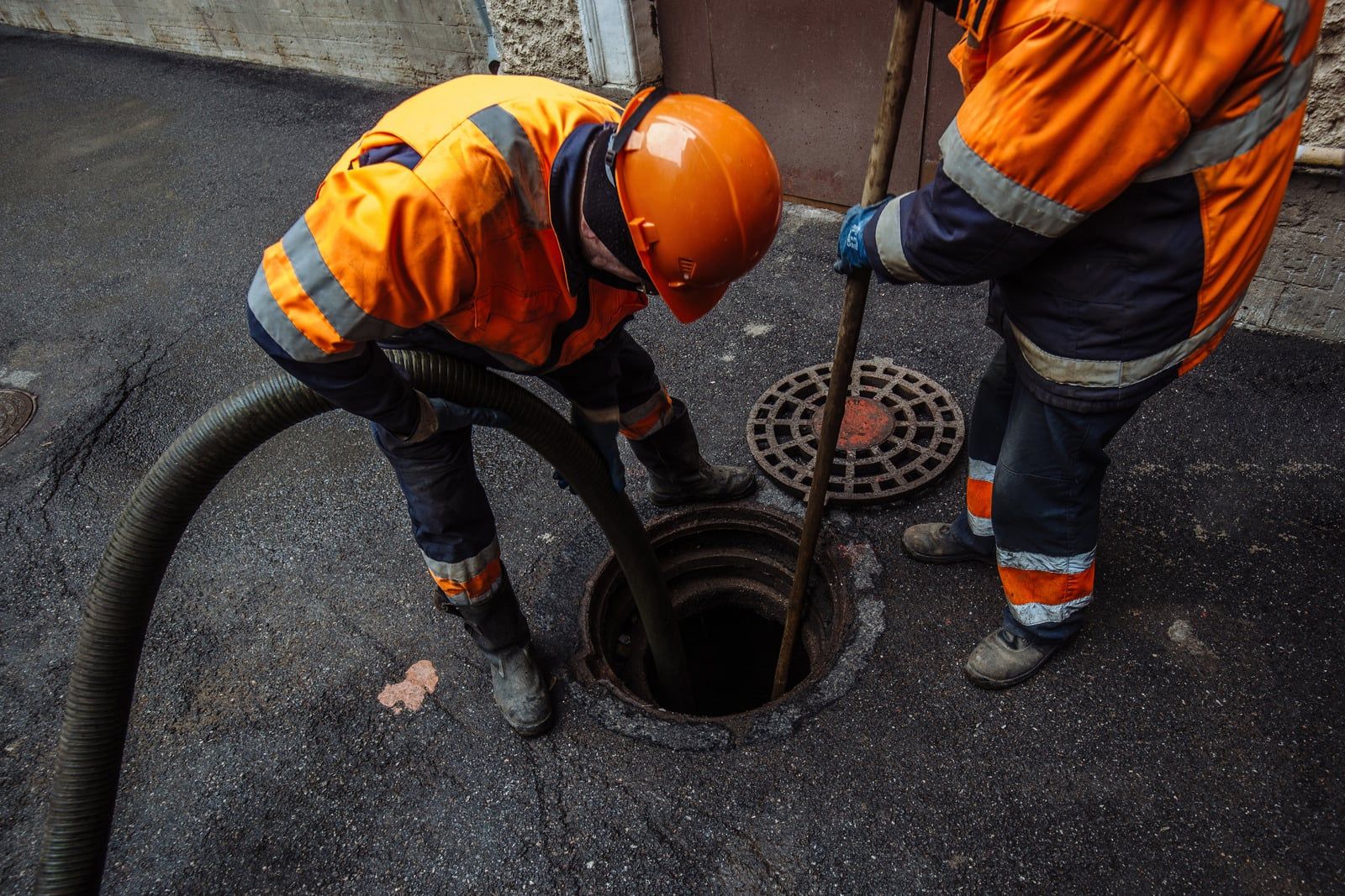 To kloakarbejdere i orange arbejdstøj spuler en kloakbrønd med en slange